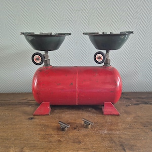 May include: A red metal propane tank with two green metal stove tops. The tank has two black knobs and two metal legs. There are two small metal knobs on the wooden surface.