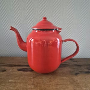 Op de afbeelding: Rode emaille theepot met een witte rand en een zwart handvat. De theepot heeft een rond lichaam en een deksel met een kleine knop.
