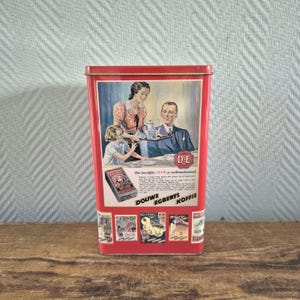 Op de afbeelding: Een vintage rode Douwe Egberts koffieblik. Het blik toont een kleurrijke illustratie van een gezin dat van koffie geniet, met de merknaam prominent weergegeven. Het blik is rechthoekig en heeft een scharnierend deksel.