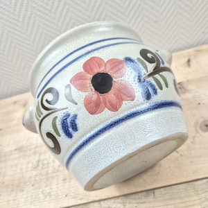 Op de afbeelding: Een witte keramische pot met blauwe en bruine beschilderde bloemenmotieven. De pot heeft twee handvatten en een licht afgeronde vorm.