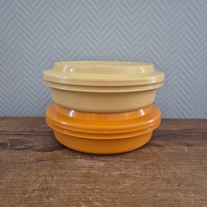 Op de afbeelding: Twee vintage plastic voedselopslagcontainers. Eén is lichtgeel van kleur en de andere is oranje. De containers zijn op elkaar gestapeld. De deksels zijn plat en passen goed op de kommen. De containers staan op een houten oppervlak.