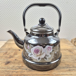 May include: A black tea kettle featuring a rose design. The kettle has a black handle and spout, and a silver chain attached to the lid. The roses are white and pink, with green leaves and stems.