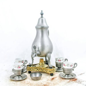 Puede incluir: Una cafetera de peltre plateada con un diseño floral en las tazas y platillos. La cafetera tiene un pico y un asa y se asienta sobre un soporte de metal dorado con tres patas. El juego incluye cuatro tazas y platillos.