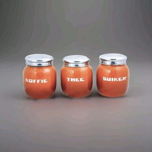 Op de afbeelding: Drie oranje keramische potten met zilveren deksels. De potten zijn gelabeld met "Koffie", "Thee" en "Suiker".
