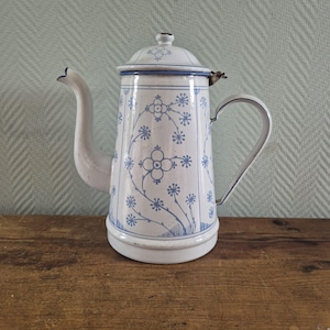Op de afbeelding: Een witte geëmailleerde koffiepot met een blauw bloemmotief van dunne, kronkelende stengels en cirkelvormige bloesems. De pot is voorzien van een gebogen tuit, een stevig handvat en een bijpassend deksel met een kleine knop aan de bovenzijde.