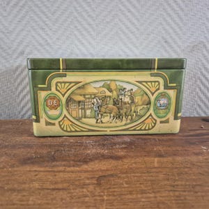Op de afbeelding: Een rechthoekig groen metalen blik met een centrale ovale illustratie van een paardenkoets voor een huis. Het ontwerp bevat goudkleurige decoratieve randen, de tekst D-E en een klein rond embleem met een afbeelding van een koe.