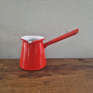 Op de afbeelding: Een felrode emaille koffiepot met een lange steel en een witte binnenkant. De pot heeft een kleine schenktuit aan de rand en een gladde, glanzende afwerking. Het ontwerp is eenvoudig en functioneel, met een cilindrisch lichaam en een steel.