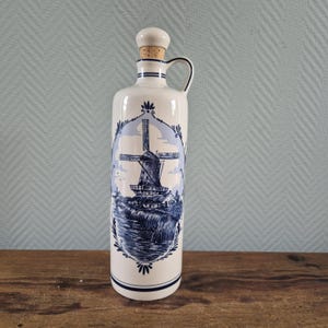 Op de afbeelding: Een witte keramische fles met een blauw windmolenontwerp. De fles heeft een handvat en een kurk. De windmolenscène is omlijst door een decoratieve rand. De fles staat op een houten oppervlak.