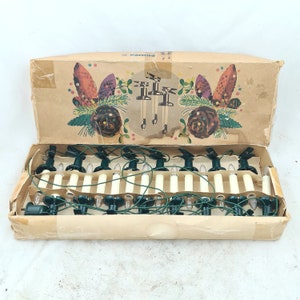 Op de afbeelding: Een doos met vintage Philips kerstverlichting met 18 witte lampjes en groene draad. De doos heeft een feestelijk ontwerp met dennenappels en hulst.