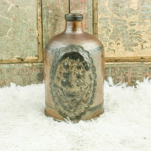 Op de afbeelding: Een bruine keramische fles met een zwart-wit ontwerp van een schip. De fles heeft een gegolfde rand rond het ontwerp en de tekst "August 1918" is zichtbaar.