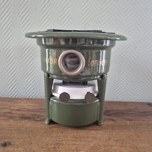Op de afbeelding: Een antieke groene Beacon Doevine kachel. De kachel heeft een ronde bovenkant met een reeks zwarte rechthoekige stukken. De kachel heeft een cirkelvormige opening met een zilveren rand. De woorden "Beacon" en "Doevine" staan op de kachel.