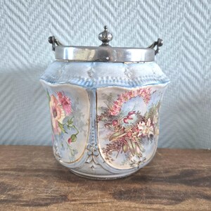 Op de afbeelding: Een vintage keramische pot met een zilveren deksel en handvat. De pot is lichtblauw met bloemmotieven in roze, rood en groen. Het deksel heeft een kleine decoratieve knop. Een decoratief stuk voor in huis.