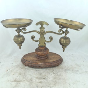 Puede incluir: Balanza antigua de latón con dos cuencos redondos y una base de madera. La balanza tiene detalles ornamentados y un diseño decorativo.