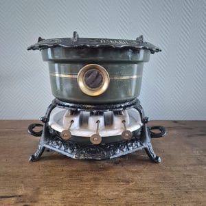 Pode incluir: Uma panela de fondue verde e preta antiga com uma base decorativa. A panela tem uma faixa dourada e um botão de cor latão. A base tem três queimadores e uma superfície de cerâmica branca. A panela está sobre uma superfície de madeira.