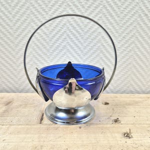 Op de afbeelding: Een kobaltblauwe glazen suikerpot met een zilveren metalen voet en een zilveren metalen handvat. De pot heeft een decoratief ontwerp van zilveren metalen bladeren rond de rand.