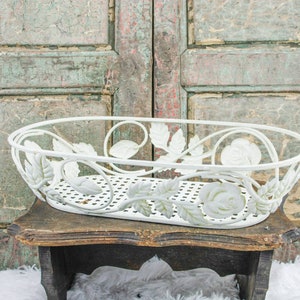 May include: White metal planter with a floral design. The planter is oval shaped and has a wire mesh bottom. It is sitting on a wooden stool.