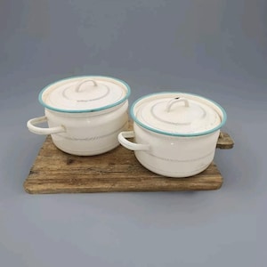 Puede incluir: Dos ollas de esmalte blanco con bordes azules y tapas. Las ollas tienen un diseño de rayas blancas y negras. Las ollas están sobre una tabla de cortar de madera.