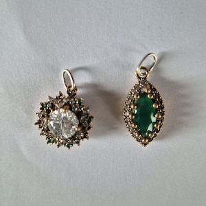 Op de afbeelding: Twee sierlijke hangers. Eén hanger heeft een grote, heldere, ronde steen omringd door kleine groene stenen en goudkleurig metaal. De andere hanger heeft een groene, ovaalvormige steen met een goudkleurige metalen zetting en kleine, heldere stenen.