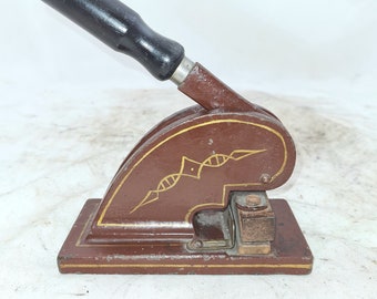 May include: A vintage brown metal stamp with a black wooden handle. The stamp has a decorative design with gold accents and a small metal lever.
