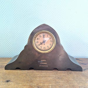 May include: Antique brown mantel clock with a round clock face and gold-colored trim. The clock has a decorative floral design on the base and a curved top. The clock face displays the numbers 1 through 12.