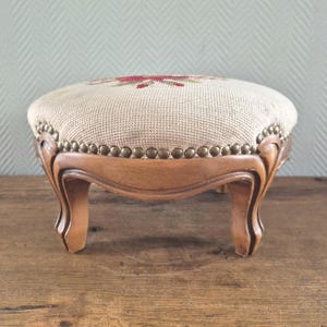 May include: A round footstool with a light beige needlepoint top featuring a red and green floral pattern. The base is crafted from medium-toned wood with curved legs and a decorative border of brass-colored metal nailhead trim along the edge.
