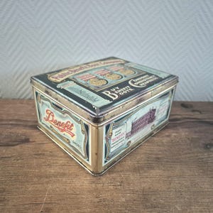 Op de afbeelding: Een vintage blikken doos met een goud en zwart ontwerp. De doos heeft de tekst "Public Benefit Shoe Company" en "Buy Benefit Boots" op de bovenkant. De doos heeft ook een afbeelding van een gebouw en de tekst "Trade Mark Benefit Shoes" aan de zijkant.
