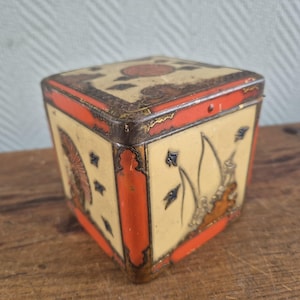 Op de afbeelding: Een antieke vierkante blikken doos met een crèmekleurige achtergrond, oranje randen en decoratieve illustraties. Het deksel heeft een centraal cirkelvormig ontwerp. De doos heeft een vintage, verouderde uitstraling.