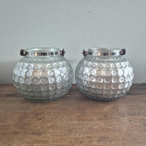 Op de afbeelding: Twee zilveren glazen kaarshouders met een getextureerd, gekarteld oppervlak en zwarte metalen handvatten.