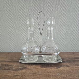 Op de afbeelding: Een vintage glazen cruetset met twee heldere, druppelvormige flessen, elk met een stopper, gehouden in een zilverkleurig metalen rek. De flessen staan op een bijpassende tray. De set staat op een houten oppervlak.