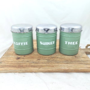 Op de afbeelding: Drie vintage groene metalen canisters met zilveren deksels, met de tekst "KOFFIE", "SUIKER" en "THEE" in witte letters. De cilindrische canisters staan op een houten oppervlak. Een charmant keuken decoratie item.