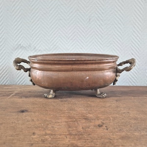 Op de afbeelding: Een ovale koperen kom met een gladde, metalen afwerking. De kom is voorzien van twee sierlijke, getextureerde messing handgrepen aan weerszijden en rust op vier kleine, gedetailleerde messing pootjes in de vorm van dierenpoten.