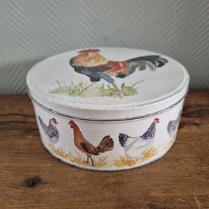 May include: A round, white metal tin with a lid featuring a rooster illustration. The sides of the tin are decorated with images of chickens. The tin sits on a wooden surface.