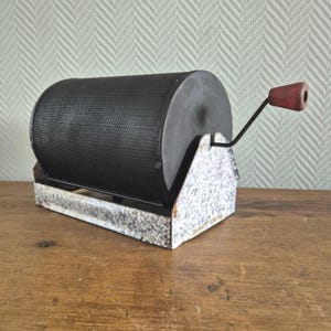 May include: An antique rock tumbler with a perforated black metal drum and a speckled white and gray base. A red wooden handle is attached to the side for turning. The tumbler is designed for polishing rocks and gemstones.