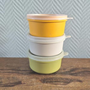 Może przedstawiać: Stos trzech małych, okrągłych pojemników do przechowywania żywności z pokrywkami. Górny pojemnik jest żółty, środkowy biały, a dolny zielony. Każdy pojemnik ma przezroczystą pokrywkę z małym uchwytem. Na drewnianej powierzchni.