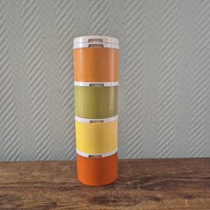 Op de afbeelding: Een stapel van vier kleurrijke, cilindervormige containers met witte deksels. De containers zijn oranje, olijfgroen, geel en een donkerder oranje. De containers zijn gestapeld op een houten oppervlak.
