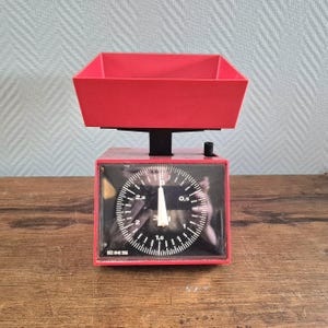 May include: A red kitchen scale with a black dial and a red bowl. The dial has markings for grams and pounds.