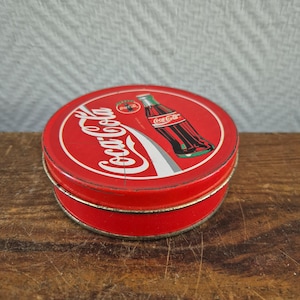 May include: A round red metal tin featuring the white Coca-Cola logo script and a graphic illustration of a glass Coca-Cola bottle. The lid displays the brand name and a small circular logo, with a visible vertical scratch across the center.