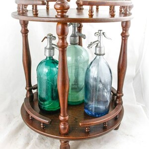 Op de afbeelding: Een houten standaard met drie glazen flessen met metalen doppen. De flessen zijn groen, blauw en helder. De standaard heeft een ronde bovenkant en drie poten.