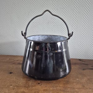 Pode incluir: Uma panela de metal esmaltado preto com um corpo largo e arredondado e uma alça de arame arqueada e resistente fixada a dois ganchos laterais. O interior da panela apresenta um acabamento salpicado de cinza e branco, contrastando com o exterior preto brilhante.