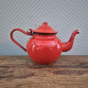 Op de afbeelding: Kleine, rode emaille theepot met een gebogen handvat en tuit. De theepot heeft een deksel en staat op een houten oppervlak. De theepot is helderrood met zwarte rand.