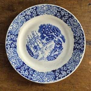 May include: A white ceramic plate with a blue floral border and a central blue illustration of a castle, tree, and figures. The plate has a vintage aesthetic, suitable for display or dining.