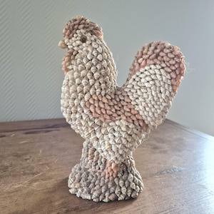 Puede incluir: Una escultura de gallo hecha completamente de conchas marinas. El gallo está de pie sobre una superficie de madera. El gallo está hecho de una variedad de conchas marinas, incluyendo conchas blancas, marrones y rosadas.