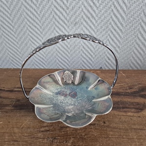 Op de afbeelding: Een zilverkleurige, geschulpte schaal met een handvat. De schaal heeft een bloemmotief op het handvat en een klein embleem in het midden. De schaal staat op een houten oppervlak.