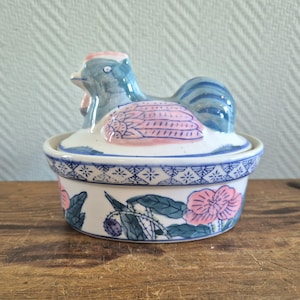 Op de afbeelding: Een keramische schotel met een deksel in de vorm van een kip, versierd met blauwe, roze en groene bloemenmotieven. Het deksel is kipvormig met een blauw en roze lichaam en een witte snavel.