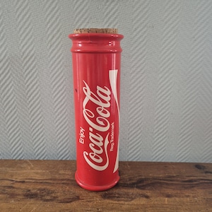 Op de afbeelding: Een rode, cilindervormige container in de vorm van een Coca-Cola-fles met een kurk. Het witte Coca-Cola-logo en de woorden "Enjoy" en "Reg. Trademark" staan op de zijkant. Het staat op een houten oppervlak.