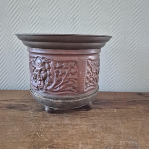 Op de afbeelding: Een koperkleurige metalen plantenbak met een gedetailleerd reliëfontwerp met een gezicht en bloemmotieven. De plantenbak heeft een brede rand, een afgeronde body en staat op drie kleine voetjes. Het ontwerp is vintage.