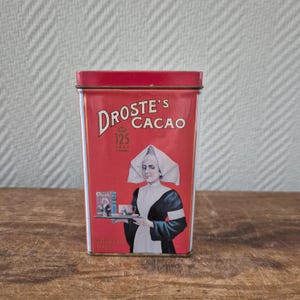 Op de afbeelding: Een vintage rode rechthoekige Droste's Cacao-blik met een rood deksel. Het blik toont de tekst "DROSTE'S CACAO" in witte letters, een vrouw met een witte kap en een kleiner beeld van een vrouw die cacao serveert. Het blik leest "125 JAAR".