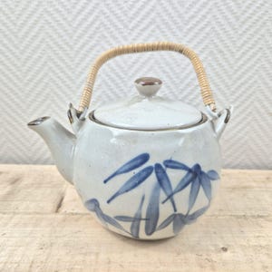 Puede incluir: Una tetera de cerámica blanca con un diseño de hojas de bambú azules y un asa tejida.
