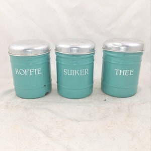 Op de afbeelding: Drie vintage teal blauwe metalen kannen met zilveren deksels. De kannen zijn gelabeld met "KOFFIE", "SUIKER" en "THEE".