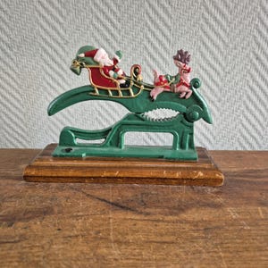 Puede incluir: Un cascanueces de metal verde montado sobre una base rectangular de madera. La parte superior del cascanueces presenta una escena pintada de Santa Claus con traje rojo en un trineo con adornos dorados tirado por un reno marrón con arnés rojo.
